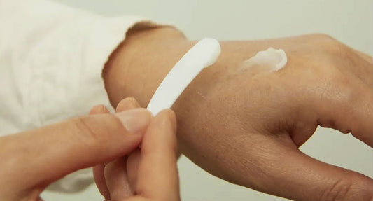 Close-up of a person applying cream to the back of their hand using a white spatula, demonstrating a skincare routine.